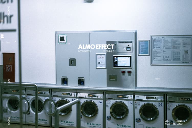 While looking through the front window of a laundromat you can see several washing machines lined up in a row. Above of them one can find the control panel of the laundromat, which receives the money for the upcoming wash cycle and can access every washing machine and dryer in the whole room. The scenery was captured by night and the dominant color of it is the blue white of the neon lights inside the shops.